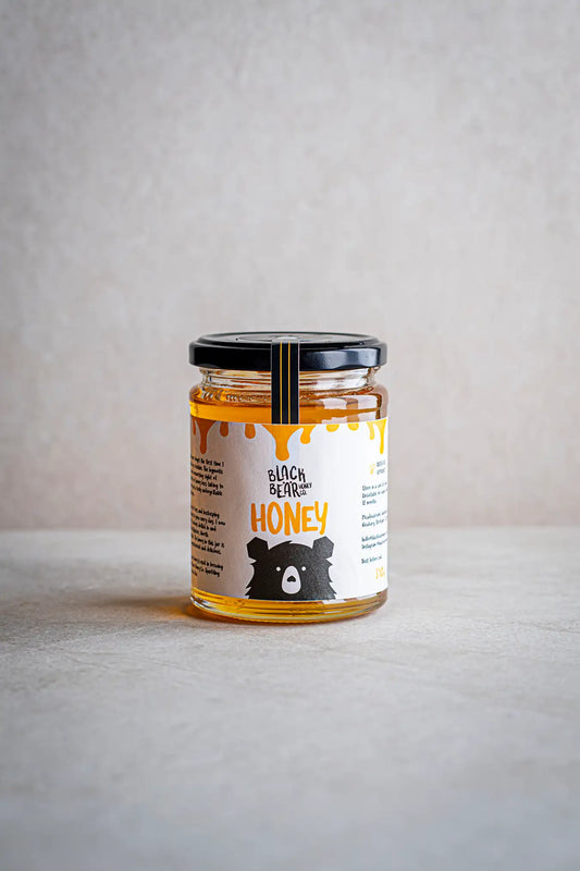 A glass jar of Honesty’s Black Bear Honey Co. Gift Pack has a black lid and a label with a cartoon bear and dripping honey design, shown on a light gray surface against a neutral background—perfect for gifting.