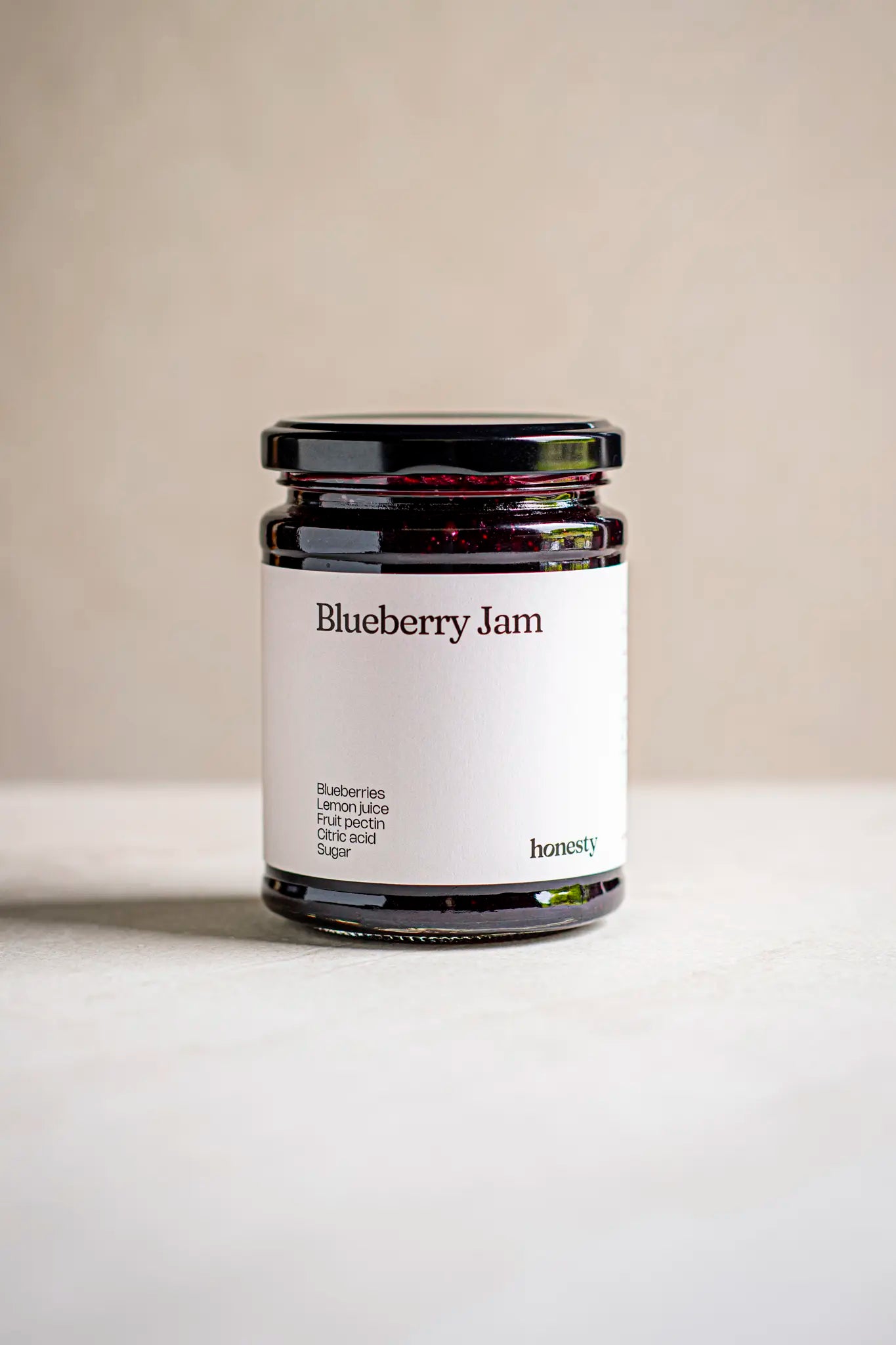 A glass jar of Honesty Blueberry Jam with a minimalist white label listing the product name and ingredients, set against a plain light background.