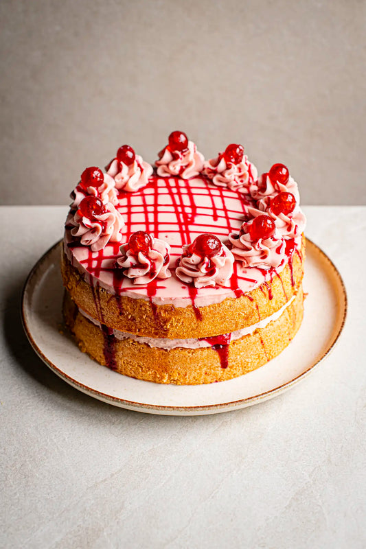 The Honesty Cherry Cake, a handmade two-layer cake with pink frosting, cream swirls, red cherries, and red syrup drizzles, is served on a white plate against a neutral background.