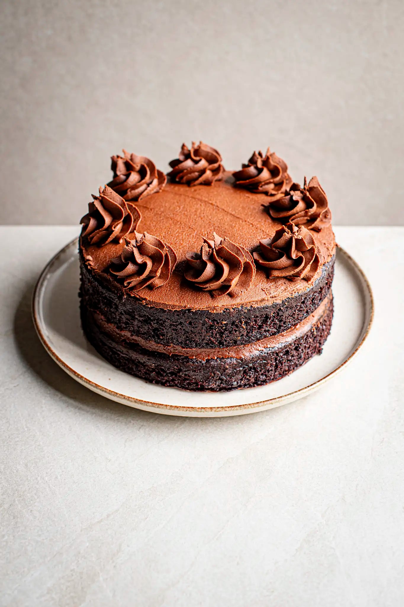 The Honesty Chocolate Cake is a handmade layer cake with eight chocolate frosting swirls, beautifully presented on a white plate against a light background—an irresistible treat.