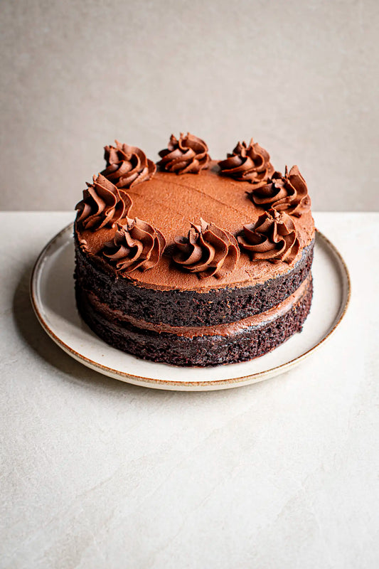 The Honesty Chocolate Cake is a handmade layer cake with eight chocolate frosting swirls, beautifully presented on a white plate against a light background—an irresistible treat.
