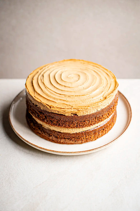The Honesty coffee cake, a three-layer naked cake with light brown frosting and a handmade spiral pattern on top, sits on a plate against a neutral background.