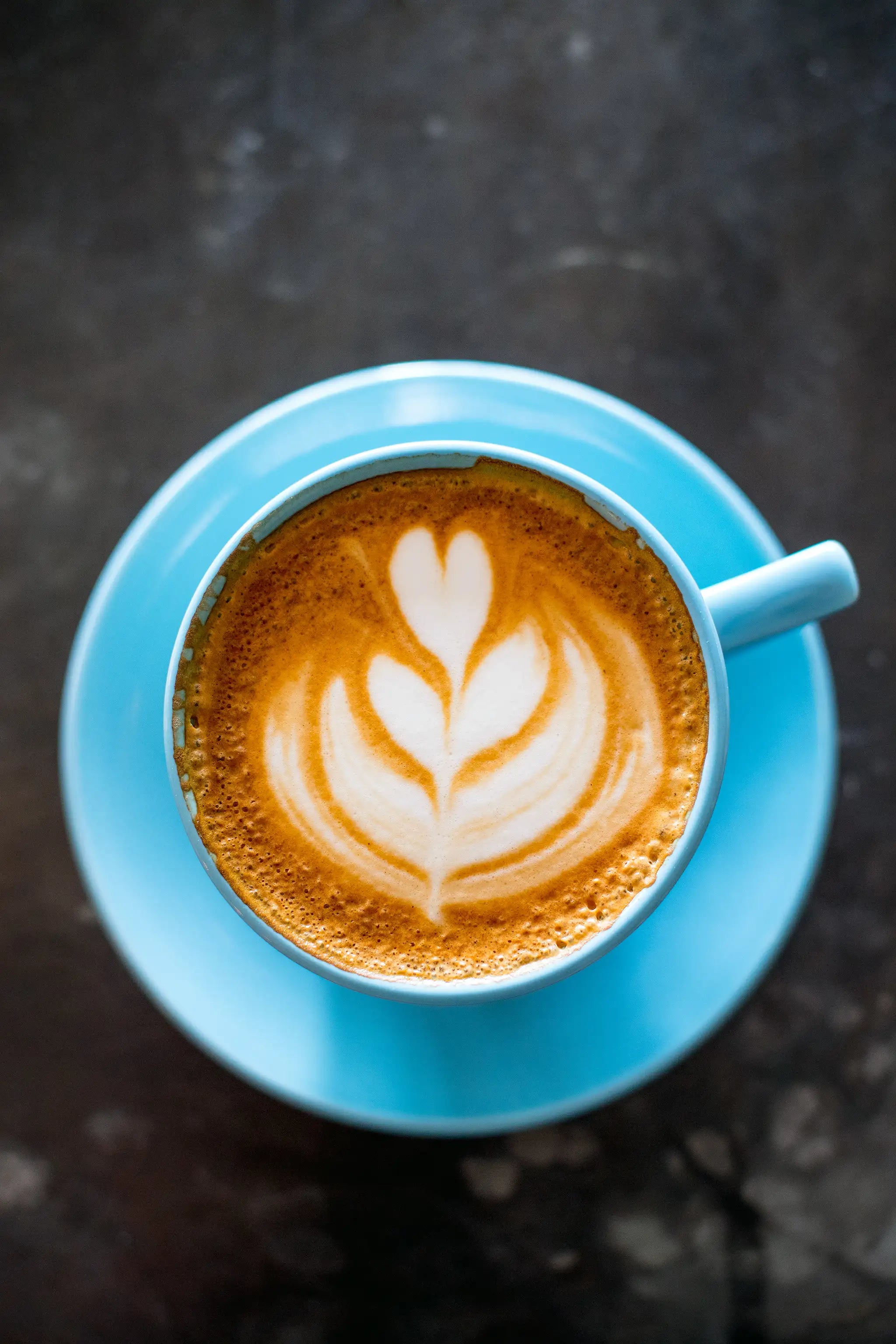 A top-down view of a cup of latte with heart-shaped latte art on the foam, served in a light blue cup and saucer on a dark surface.