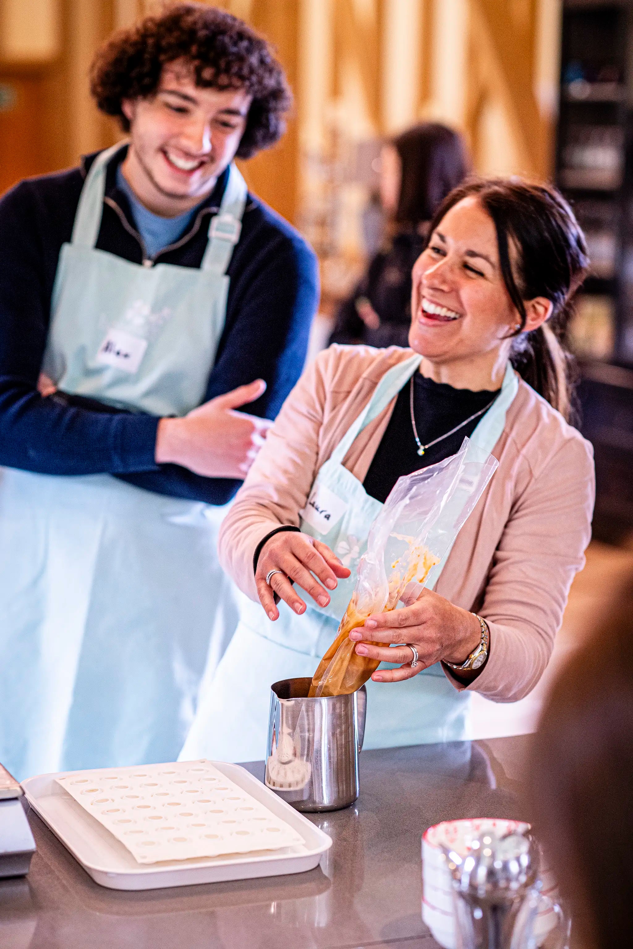 Two people wearing aprons smile and laugh together as one of them pipes a mixture into a mold in a kitchen or workshop setting.