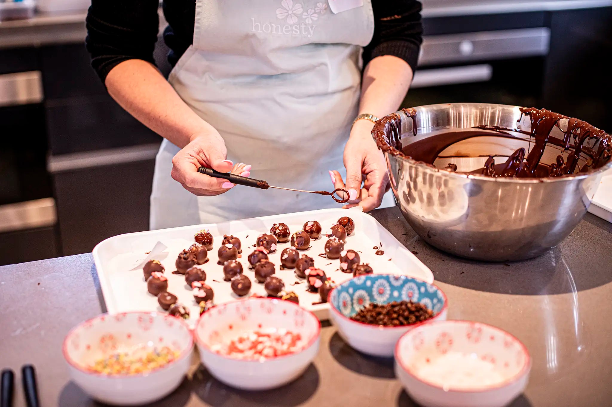 A person in an apron dips treats into melted chocolate and places them on a tray, surrounded by bowls of toppings and a large bowl of chocolate in a kitchen.