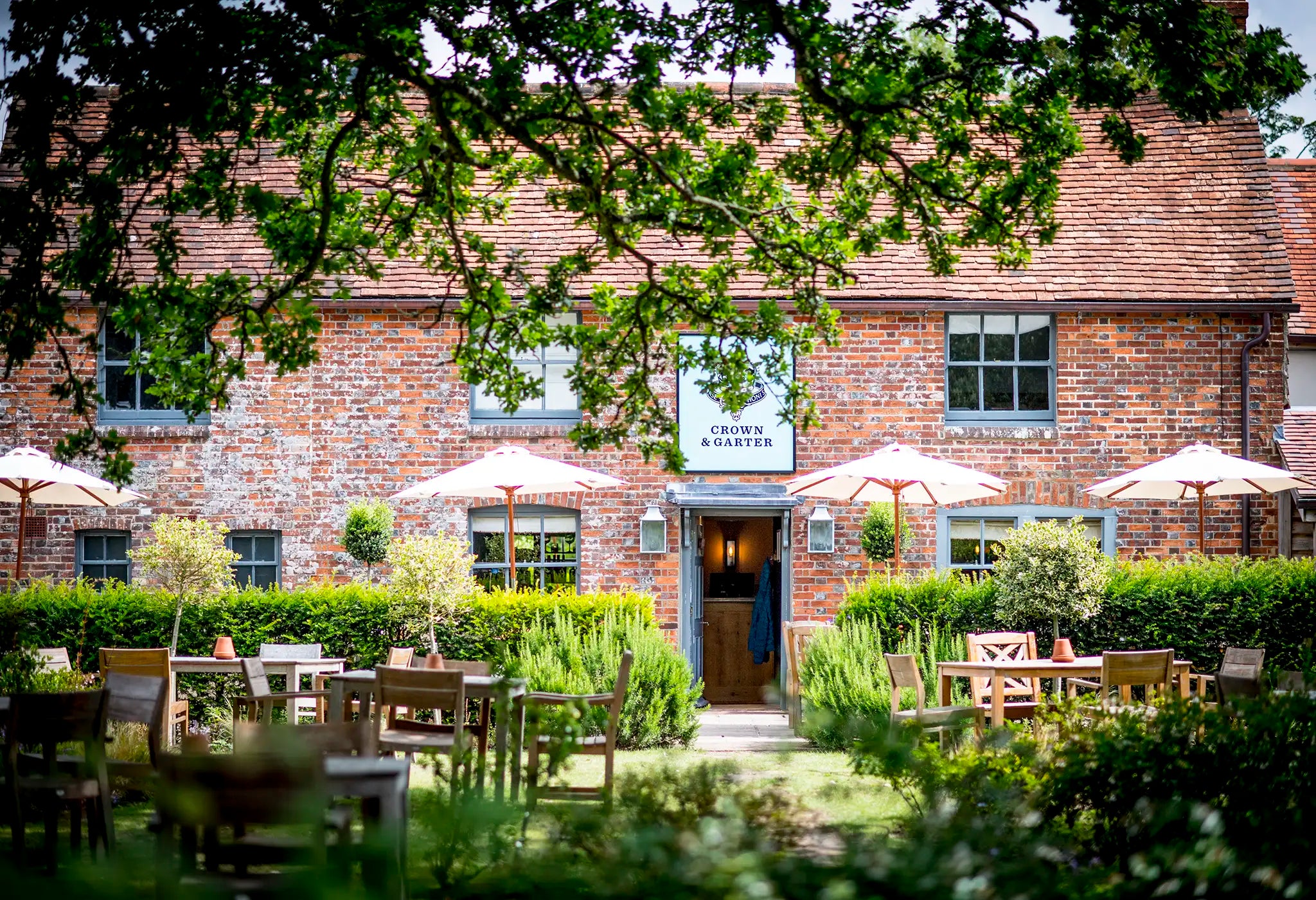 A charming brick pub with a sign reading Crown & Garter, surrounded by greenery and outdoor wooden tables and chairs under white umbrellas, framed by leafy tree branches.