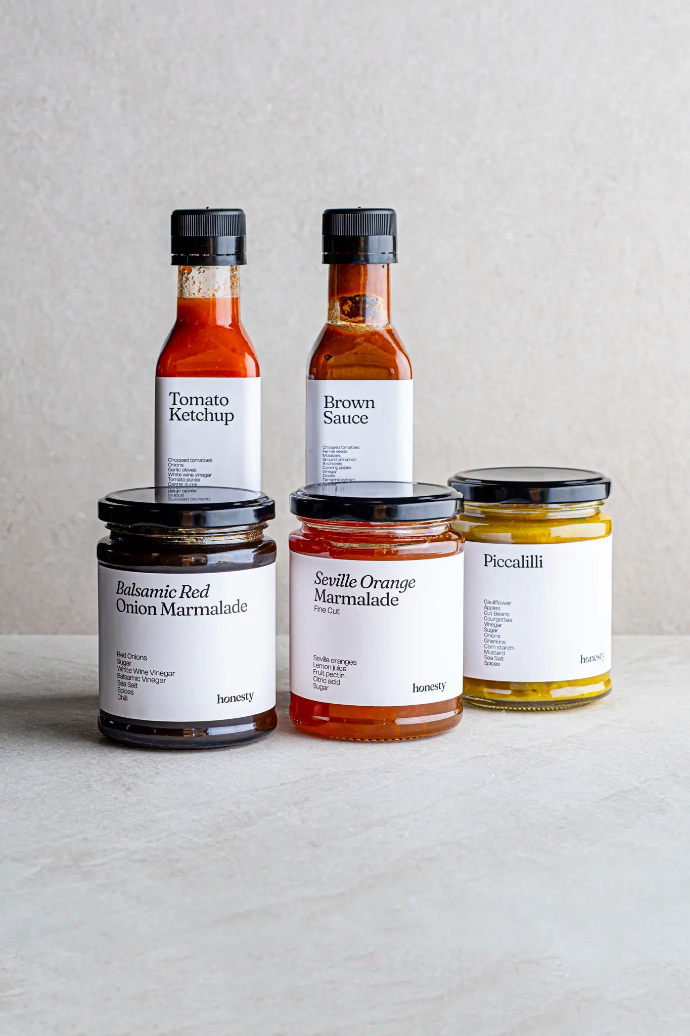 Five Honesty Cupboard Filler condiment jars and bottles are arranged on a light surface, featuring Tomato Ketchup, Brown Sauce, Balsamic Red Onion Marmalade, Seville Orange Marmalade jam, and Piccalilli.