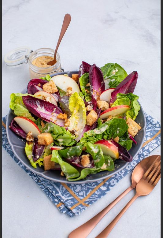 A vibrant salad of mixed greens, apples, croutons, and walnuts sits with a jar of dressing and copper cutlery—the ideal bite before enjoying the I Cant Believe its Vegan Event for those interested in plant-based cooking.