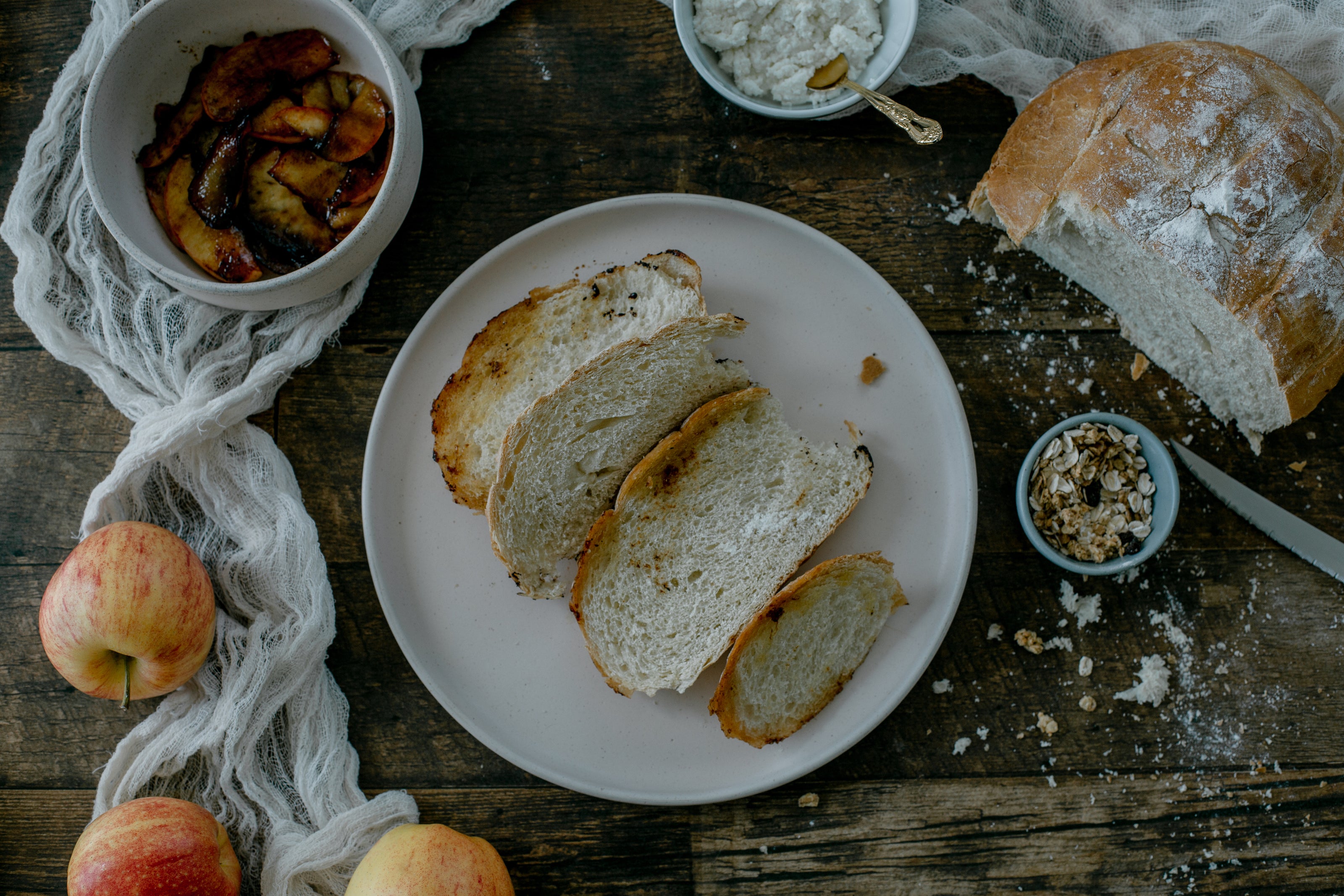 A plate with four slices of rustic bread sits on a wooden table, surrounded by a loaf of bread, a bowl of caramelized apples, whole apples, a bowl of crumbled cheese, and a small dish of granola.