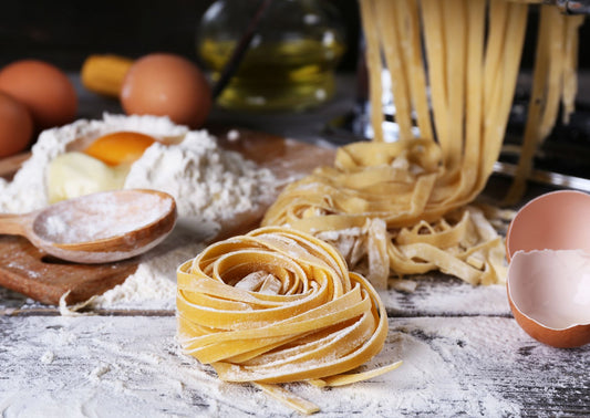At the Event’s Pasta Workshop: Fresh Tagliatelle and Neapolitan Lasagne to take home, homemade pasta nests rest on a floured surface with cracked eggs, flour, a wooden spoon, pasta machine, egg shell, and olive oil in the background.