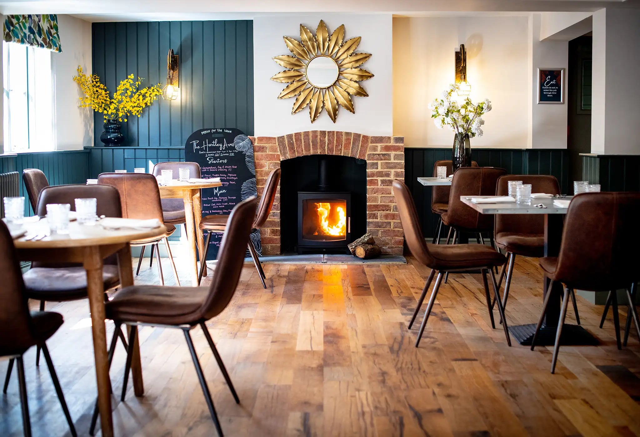 Cozy restaurant interior with wooden floors, brown chairs, and round tables. A lit fireplace with a brick surround sits beneath a sun-shaped mirror, with decorative flowers and soft lighting completing the warm, inviting atmosphere.
