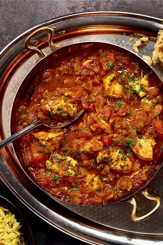 Event’s Punjabi Comforts, Flavours of Home features chicken curry in a rich red sauce with herbs, served in a round metal dish with naan and yellow rice on a silver tray. A spoon rests in the curry, inspired by Punjabi cuisine.