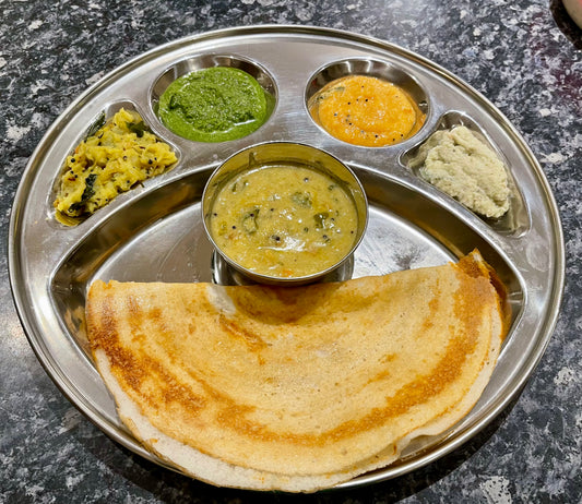 Enjoy the Pancake Day Special - Dosa Masterclass by Event: a stainless steel plate with crispy dosa, sambar, three colorful chutneys, and potato sabzi on a dark speckled countertop.