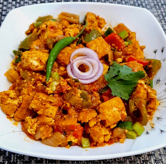 A square white plate of colorful paneer bhurji from Events Flavours of India, garnished with red onion rings, a green chili, and cilantro, is served on a woven placemat.