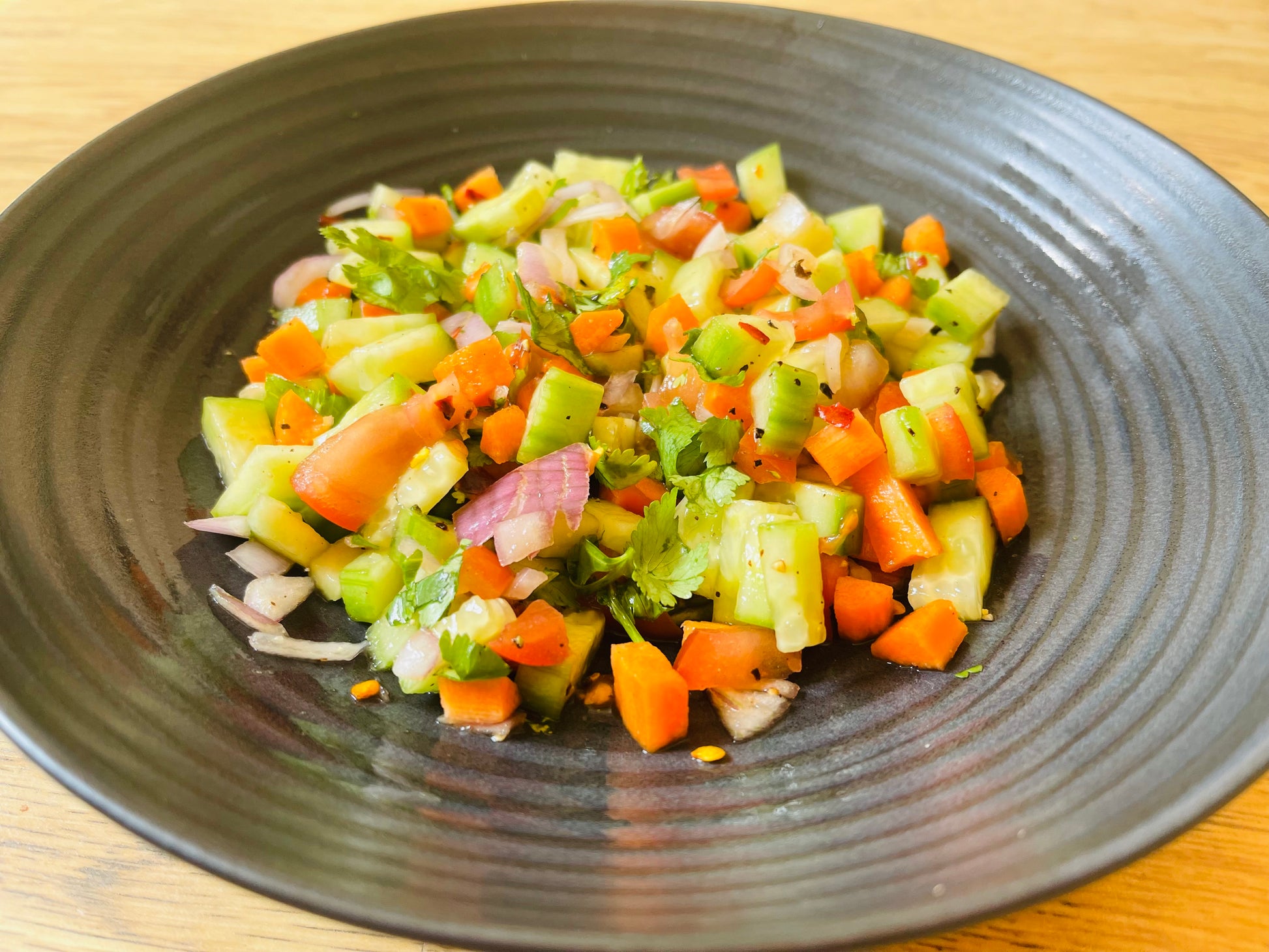 A black plate with a colorful salad of chopped cucumber, tomato, carrot, red onion, and cilantro seasoned with pepper from Flavours of India by Event captures authentic Indian taste on a wooden surface.