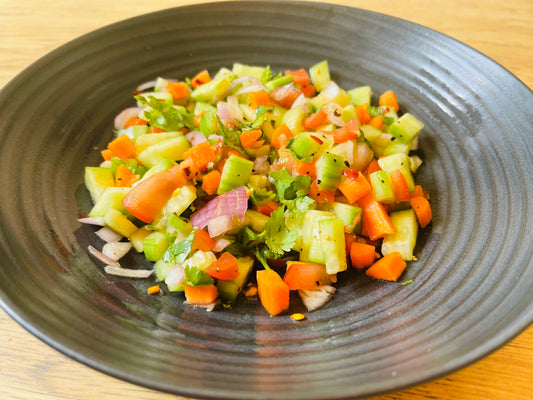 A black plate with a colorful salad of chopped cucumber, tomato, carrot, red onion, and cilantro seasoned with pepper from Flavours of India by Event captures authentic Indian taste on a wooden surface.