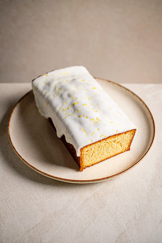 The Honesty Victoria Sponge, a handmade rectangular loaf with white icing and lemon zest, is displayed on a round beige plate atop a light surface. One end is sliced open to show the cake’s golden interior.