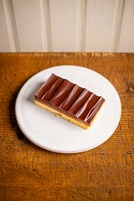 A rectangular Honesty millionaires shortbread with a glossy, wavy top sits on a white plate atop rustic wood, showing off its visible creamy and cake layers.