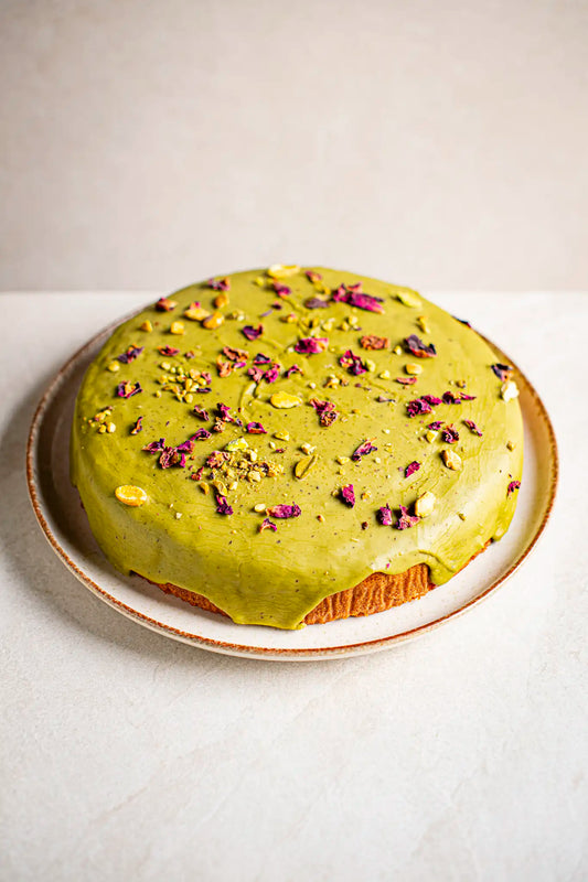 A round handmade pistachio cake by Honesty, topped with green icing, chopped pistachios, and dried rose petals, rests on a ceramic plate against a light background.