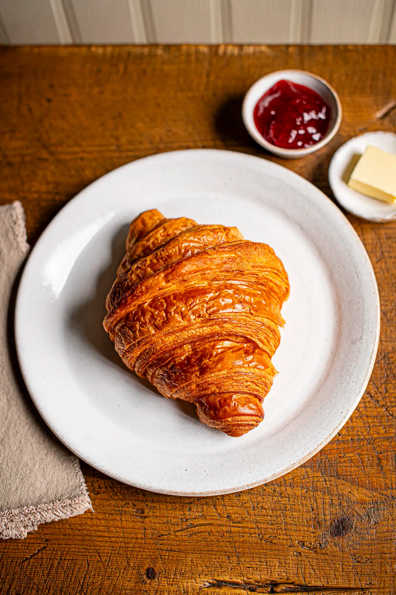 A golden Honesty Croissant rests on a white plate atop a wooden table, accompanied by a small dish of red jam, butter slices on another plate, and a beige napkin.