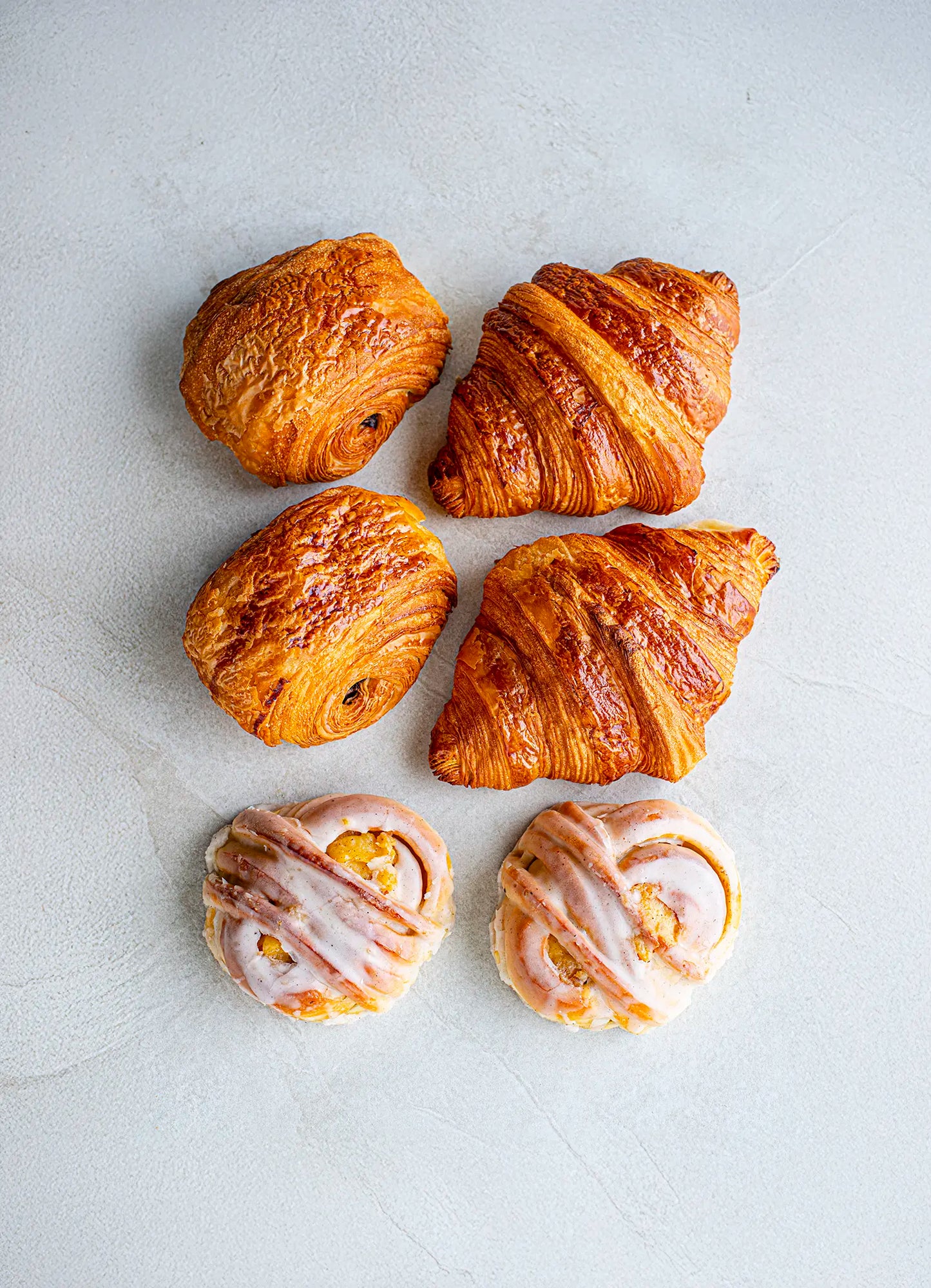Four Honesty Quick Meeting plain croissants and two Madagascan vanilla iced buns are neatly arranged on a light textured surface, with the croissants placed above the buns.