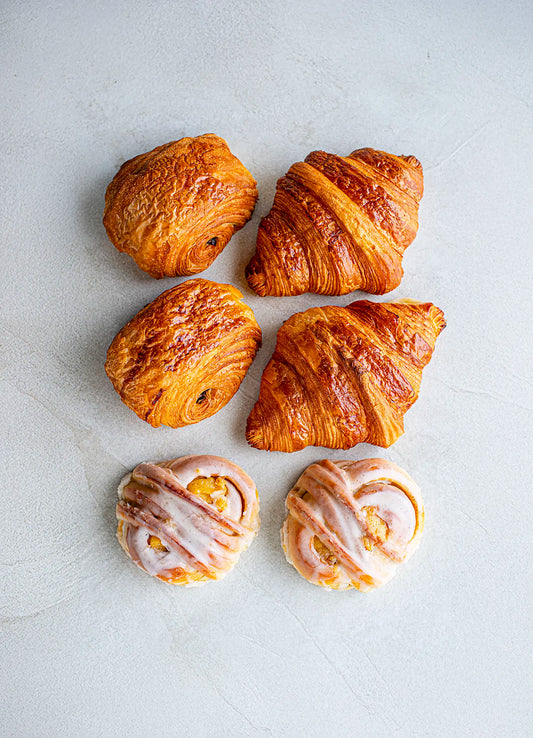 Four Honesty Quick Meeting plain croissants and two Madagascan vanilla iced buns are neatly arranged on a light textured surface, with the croissants placed above the buns.
