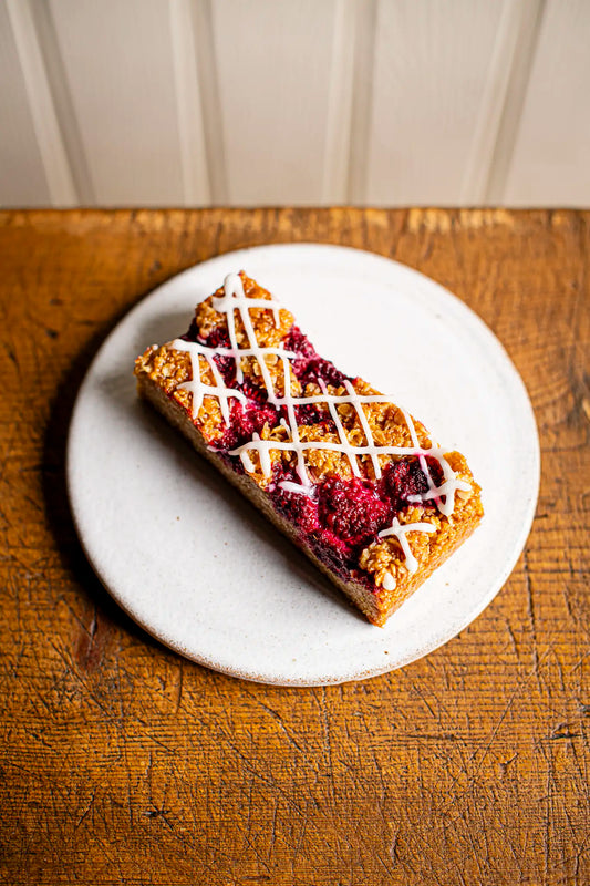 A handmade Victoria Sponge by Honesty, topped with raspberry filling and white icing drizzle, sits on a round white plate on a rustic wooden surface.