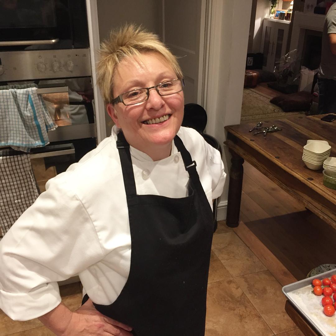 A smiling person in glasses, a chef’s coat, and black apron stands in a kitchen by cherry tomatoes and bowls, ready to share healthy recipes at the Chinese Cookery Class by Event.