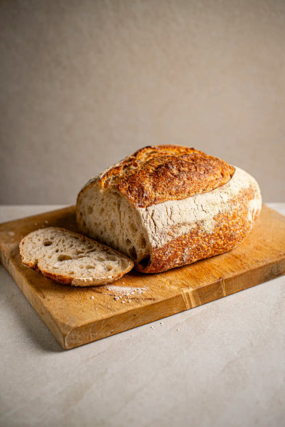A classic sourdough by Honesty sits on a wooden cutting board, with one slice cut and placed beside it. The background is simple and neutral.