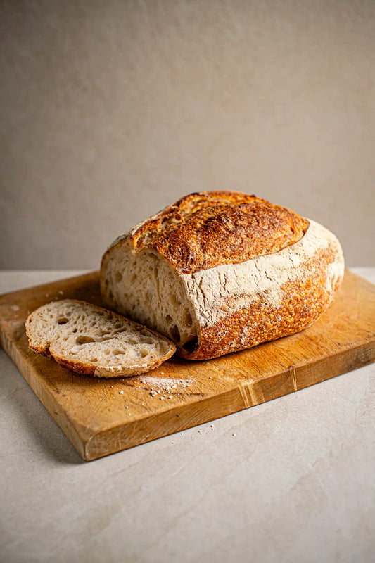 A classic sourdough by Honesty sits on a wooden cutting board, with one slice cut and placed beside it. The background is simple and neutral.