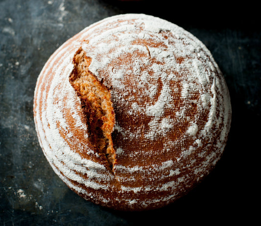 Sourdough Made Easy by Event is a round, rustic loaf with a crusty, flour-dusted surface and a deep crack on top, shown against a dark background.