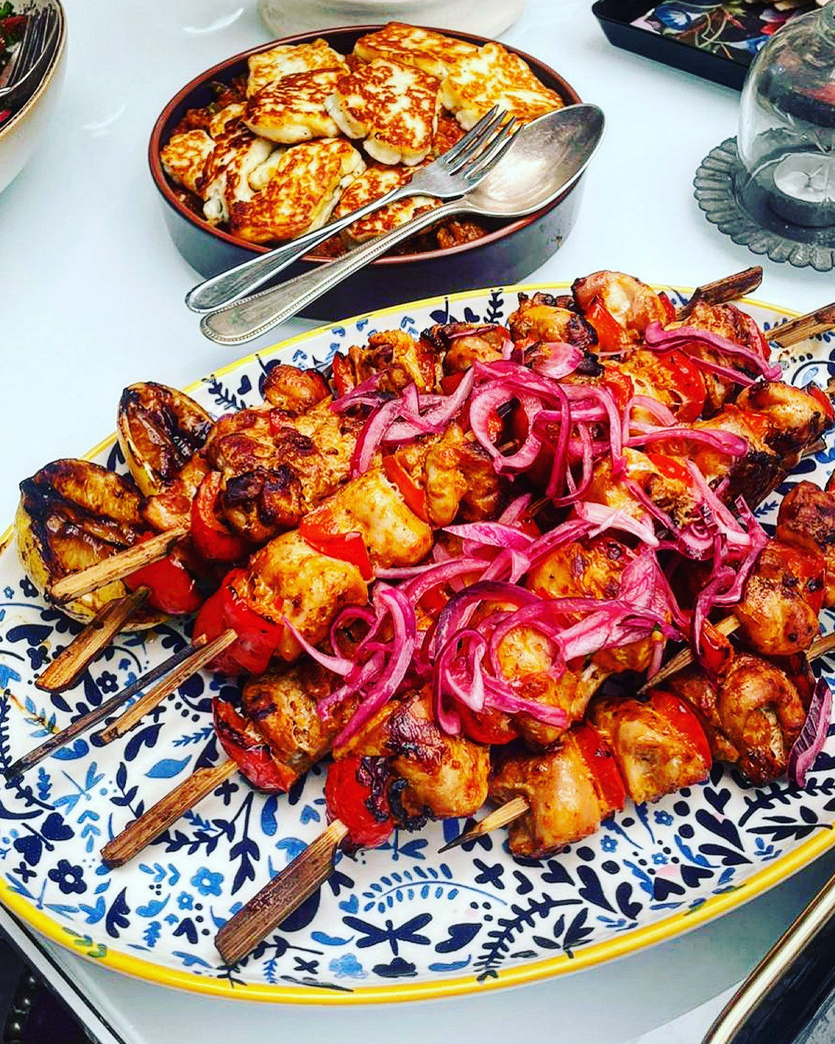 A platter from Event's The Greek Kitchen features grilled chicken and vegetable skewers with sliced red onions, served on a patterned plate. In the background, grilled cheese slices are presented with serving utensils.