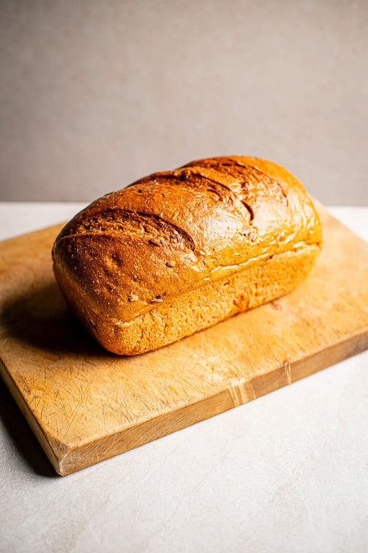 The Honesty Wholemeal Farmhouse loaf, hand baked with a golden crust, rests on a wooden cutting board, highlighting its artisanal quality against a light background.
