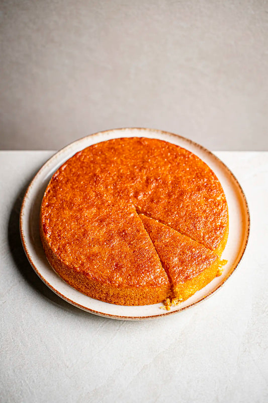 A round, golden-brown Honesty Tunisian Orange cake sits on a plate with one slice slightly pulled out, showing its moist texture. The plain cake has a shiny glaze on top and is set against a light background.