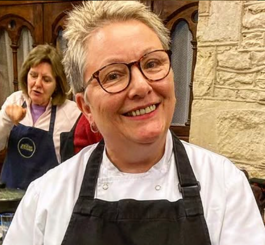 A smiling woman in a white chef’s jacket and black apron leads the Surviving Uni - Student Cookery Class by Event, guiding students indoors while another participant in a blue apron appears blurred in the background.
