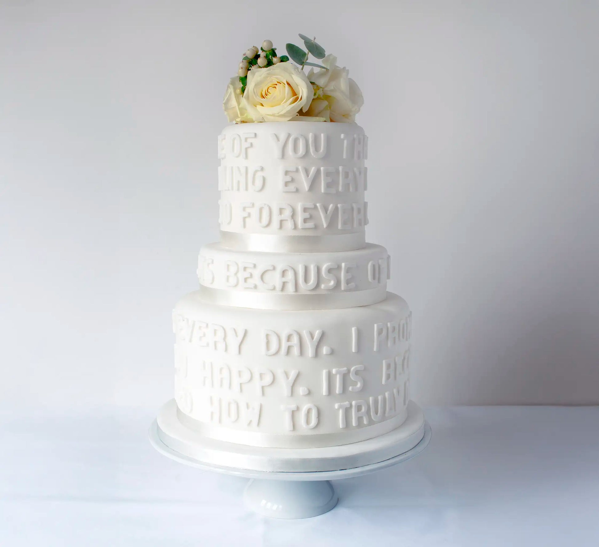 The Honesty Celebration Cake features three white tiers with embossed text, topped with white roses and greenery, making it an elegant centerpiece for any celebration. Displayed on a white pedestal against a plain background.