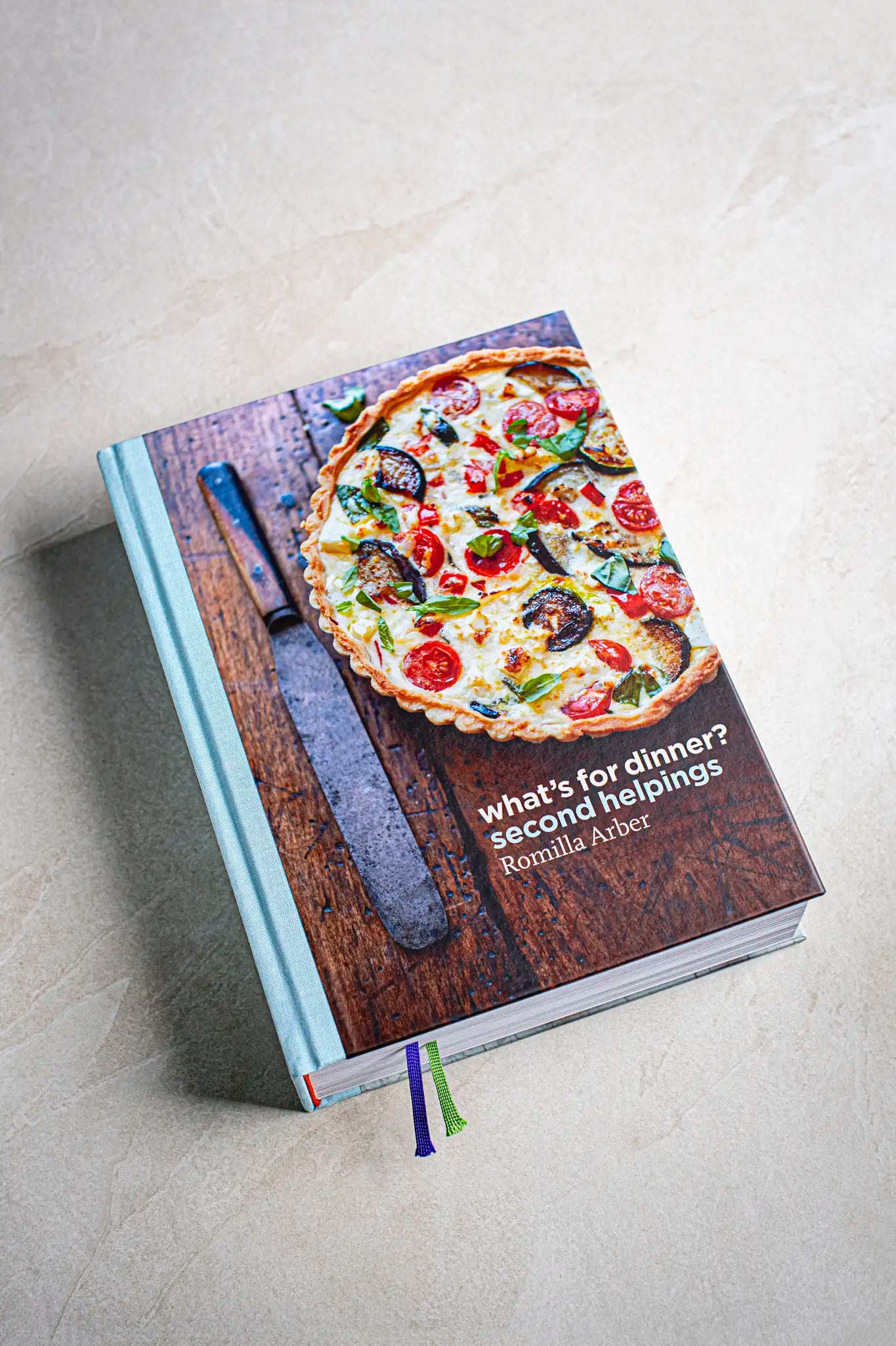 A handmade cookbook titled Victoria Sponge by Honesty, featuring a photo of a vegetable tart on the cover, rests on a light surface next to a large knife—perfect for home chefs.