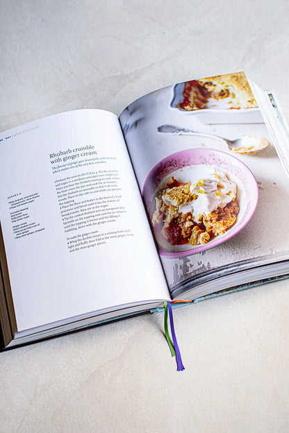 An open cookbook shows Honesty’s Victoria Sponge recipe on one page and a photo of the cake topped with cream on the opposite page, set on a light surface for an inviting display.