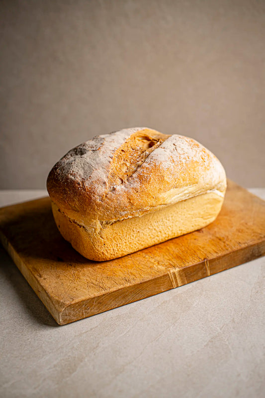 A fresh loaf of Honesty’s White Farmhouse bread with a golden crust rests on a wooden cutting board against a neutral background, highlighting its artisanal quality.